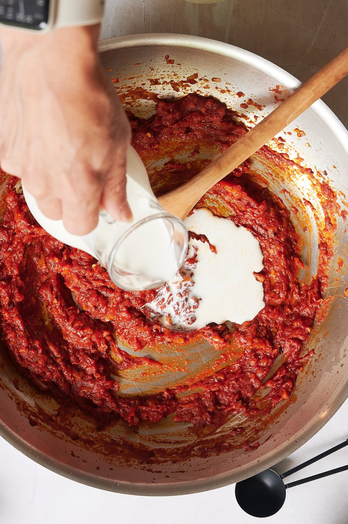 A hand pours cream from a glass bottle into a pan of spicy vodka sauce being stirred gently with a wooden spoon.