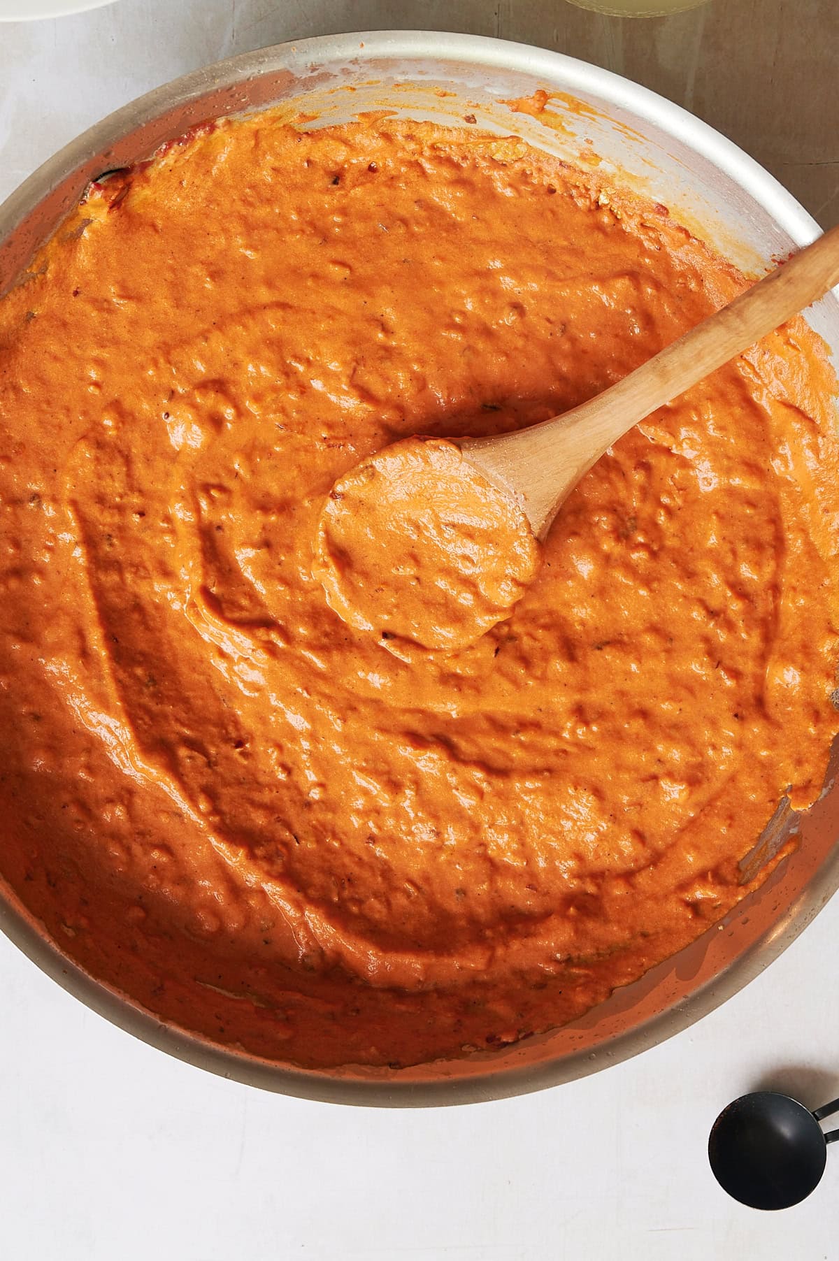A wooden spoon stirs a thick, orange-red vodka sauce in a large metal pan on a white surface.