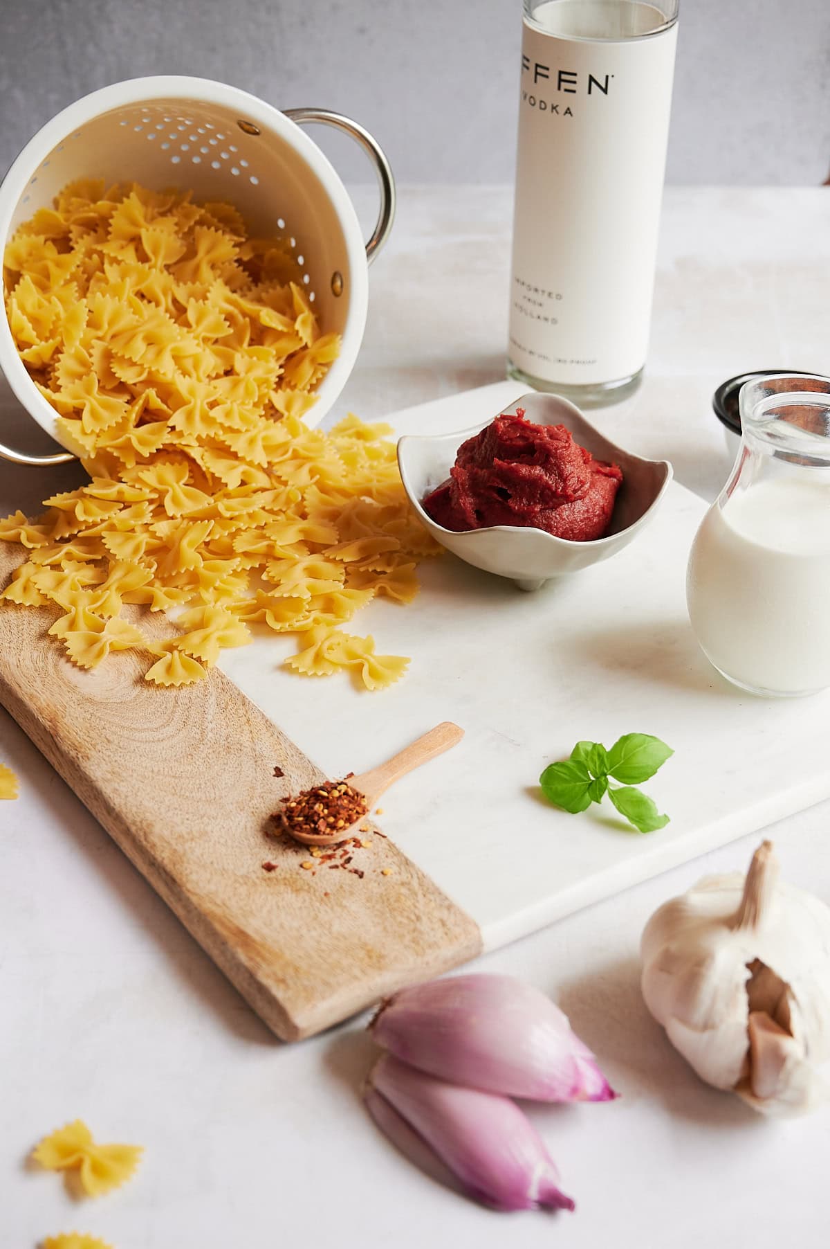 Dry bowtie pasta spills from a colander near ingredients for spicy vodka sauce with pancetta—tomato paste, cream, basil, garlic, shallots, and red pepper flakes.