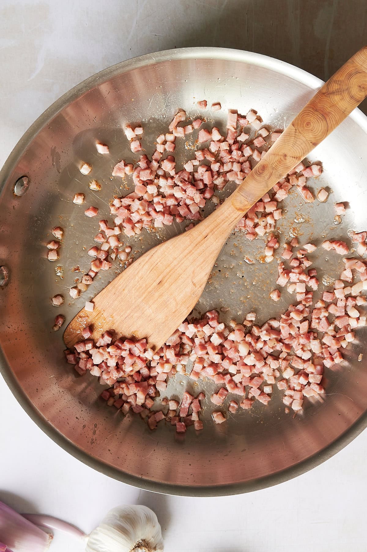 Chopped bacon pieces being sautéed in a stainless steel pan with a wooden spatula, ready to elevate a spicy vodka sauce with pancetta. Garlic and shallots rest nearby.