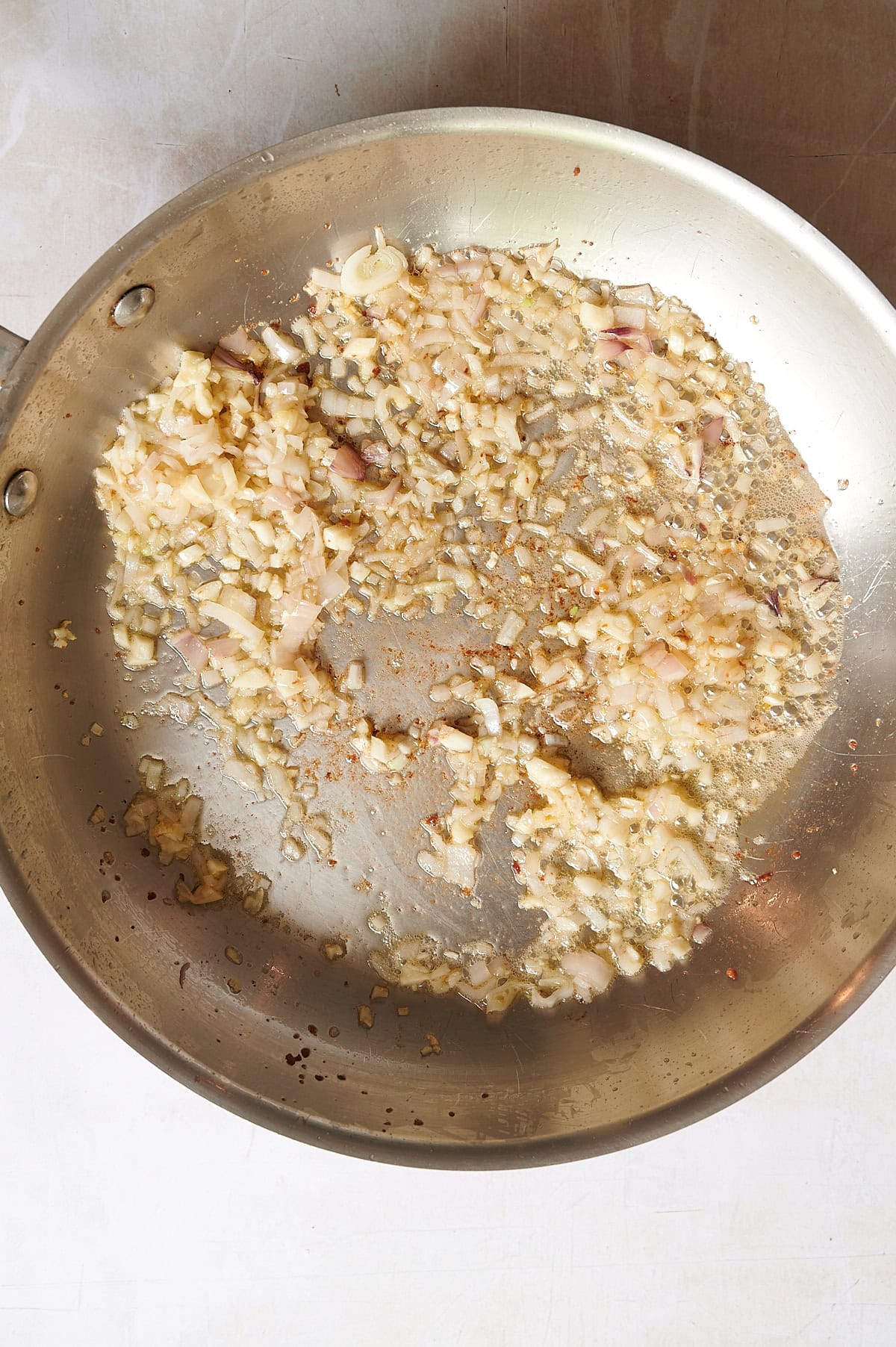 Chopped shallots and garlic sauteeing for a spicy vodka sauce with pancetta in a stainless steel pan on a light-colored surface.