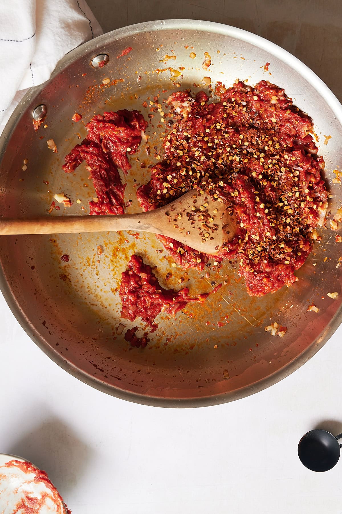 A metal pan on a white surface contains spicy vodka sauce with pancetta, tomato paste, onions, and red pepper flakes being stirred with a wooden spoon.