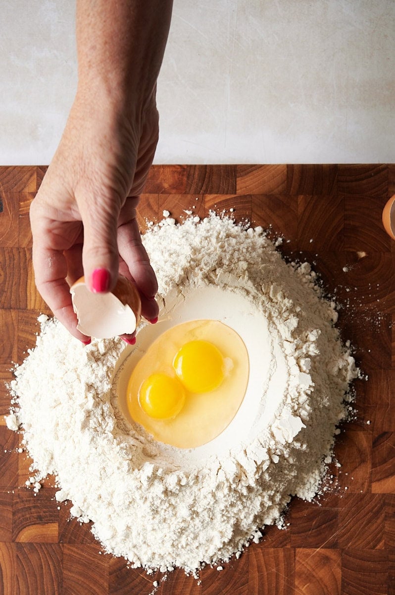 A hand cracking an egg shell over a pile of flour with two eggs in the center, on a wooden surface—perfect for making homemade ravioli dough.