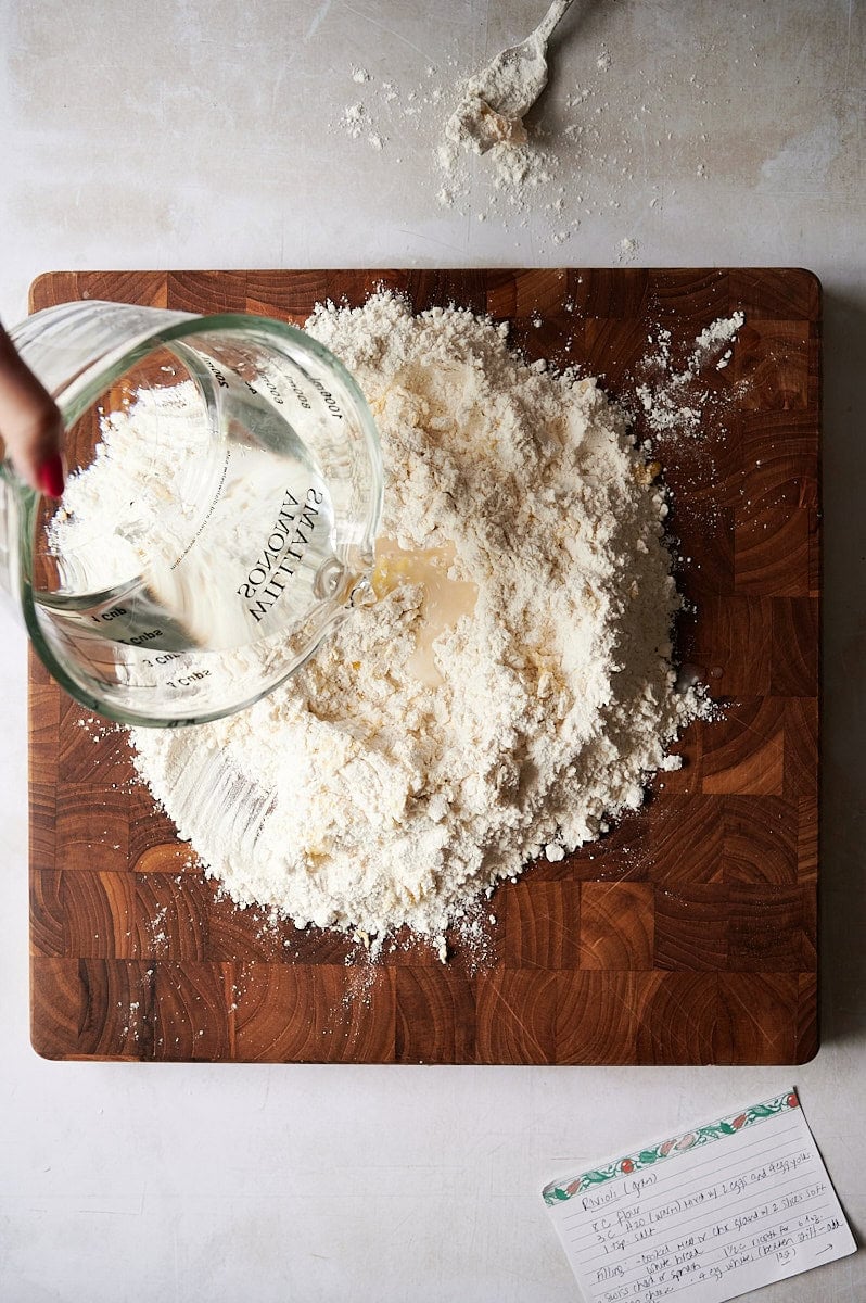 A hand pours water from a glass measuring cup into a mound of flour on a wooden board, starting homemade ravioli dough; a fork and a recipe card rest nearby.