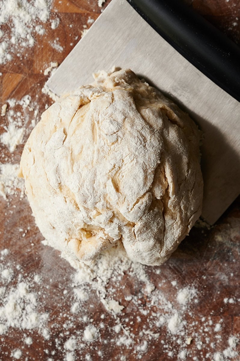 A ball of homemade ravioli dough sits on a floured wooden surface next to a metal bench scraper with a black handle.