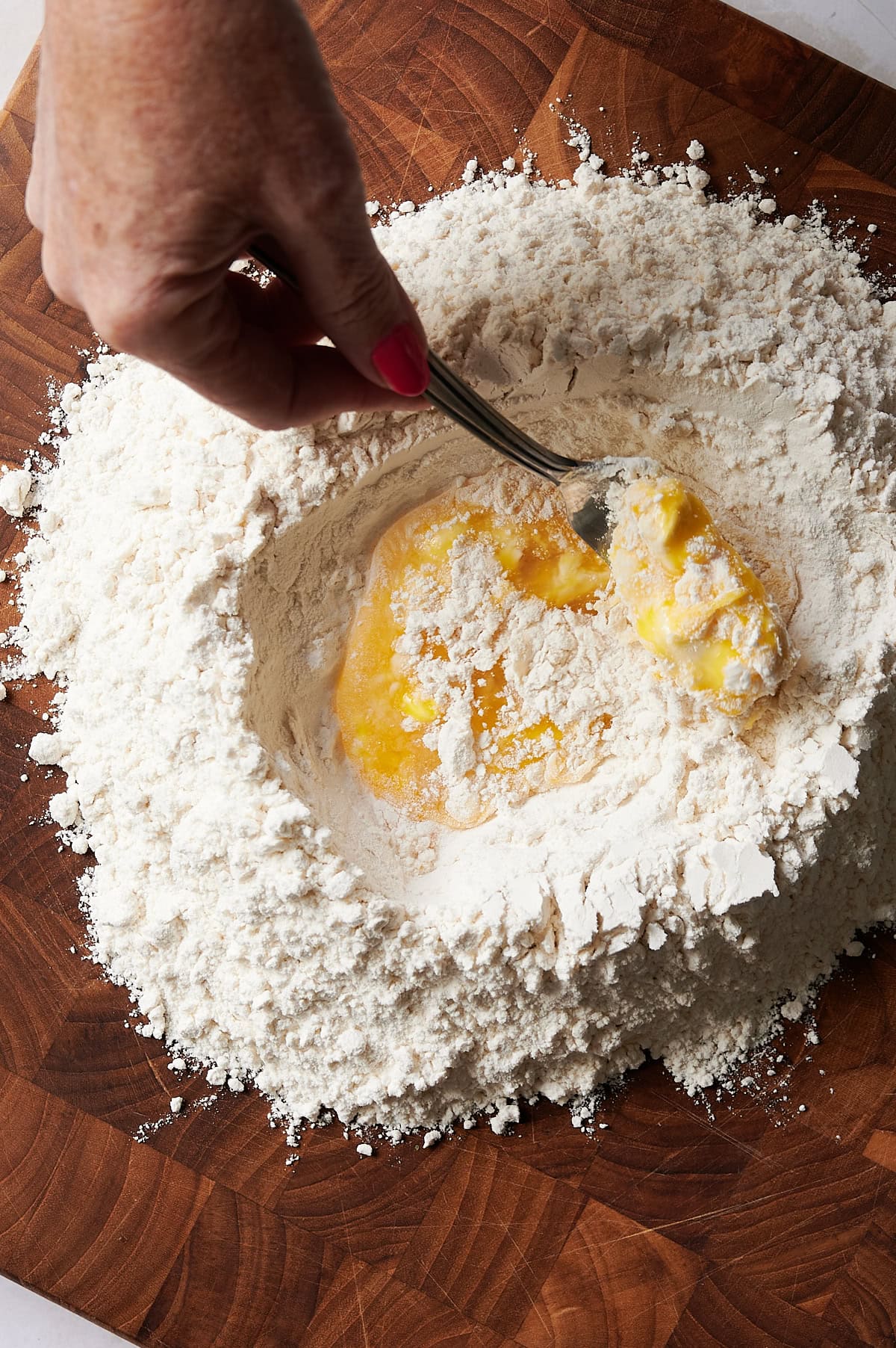 A hand uses a fork to mix eggs into a well of flour on a wooden surface, preparing homemade ravioli dough.