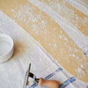rolled pasta dough strips on a blue and white dish towel with a ravioli stamp and bowl of water.