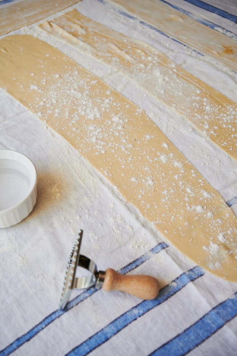 rolled pasta dough strips on a blue and white dish towel with a ravioli stamp and bowl of water.
