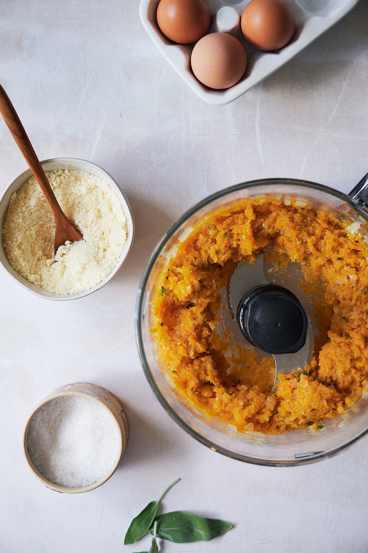 A food processor with a butternut squash ravioli filling mixture sits on a counter next to a bowl of grated cheese, a container of salt, a few sage leaves, and a carton holding three brown eggs.