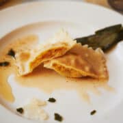 close up of a butternut squash ravioli cut in half with sage on a white plate.