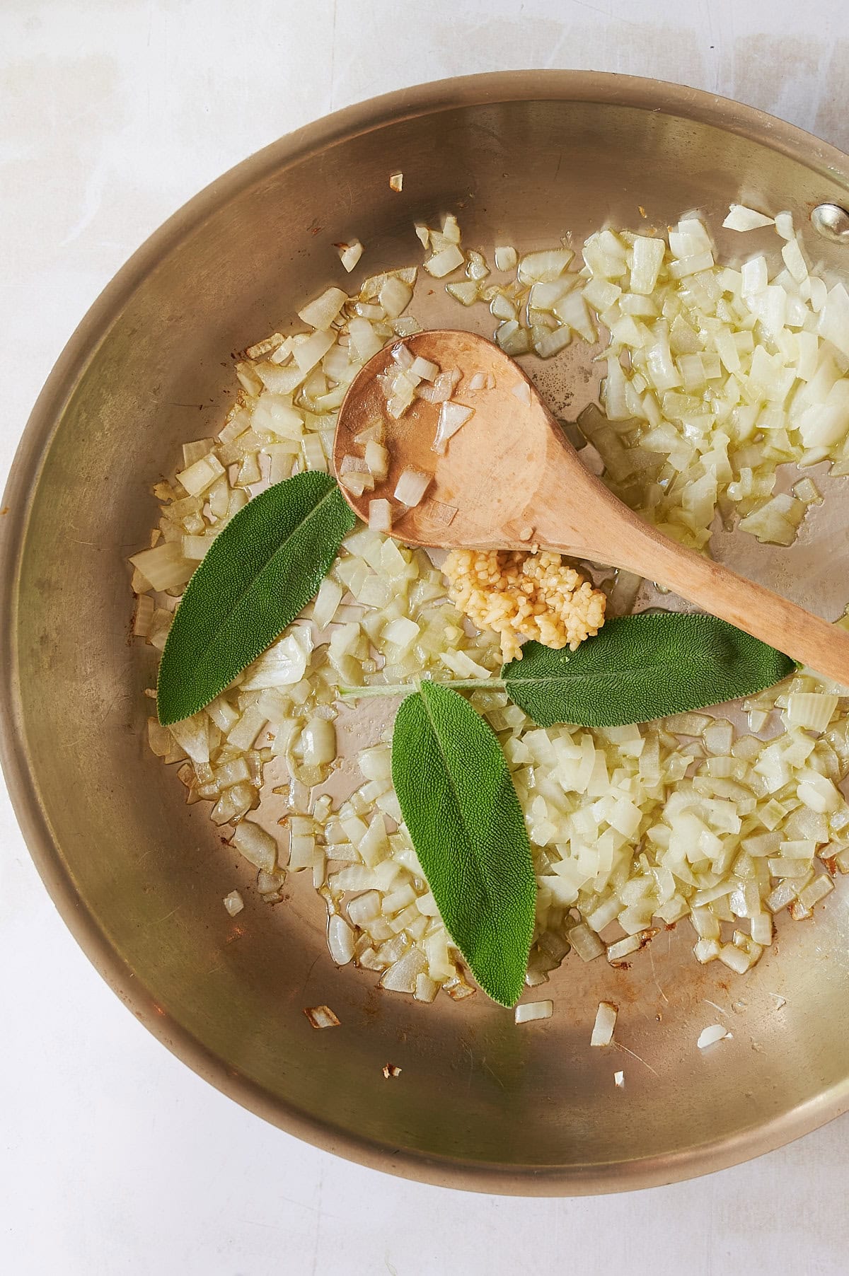 A metal pan with chopped onions, whole sage leaves, and minced garlic being sautéed for butternut squash ravioli. A wooden spoon rests in the pan as the ingredients are lightly cooked and spread out across the surface.