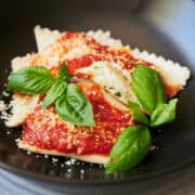 black dish with tomato sauce covered cheese ravioli and fresh basil leaves.