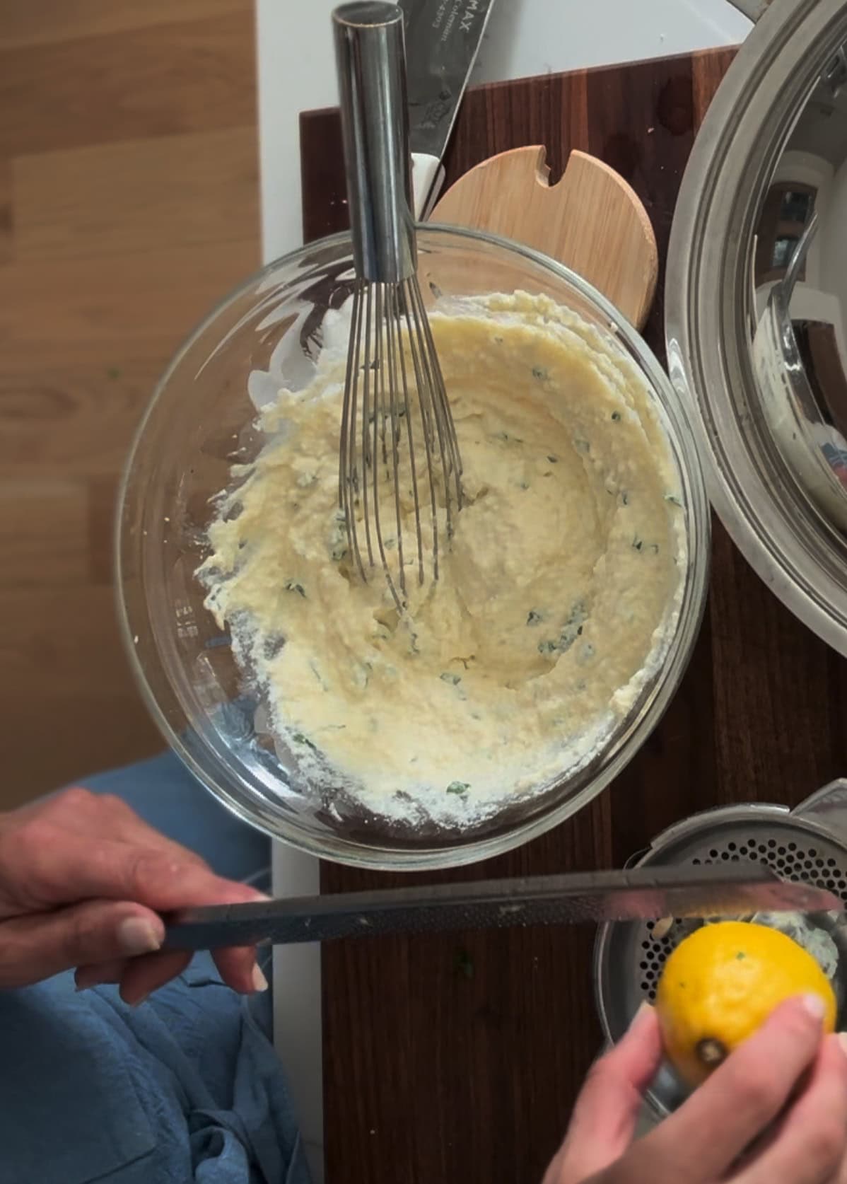 A person grates a lemon over a glass bowl of batter with herbs, using a microplane; a whisk rests in the bowl on a wooden cutting board, for preparing lasagna with ricotta.
