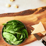 a wooden board with basil, parmesan cheese and a knife near a bowl of walnuts, olive oil bottle and garlic.