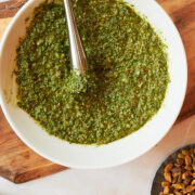 white bowl of pesto with a black muddler sitting on wooden board with bowl of pistachios