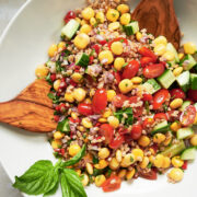 white bowl with cucumbers, lupini beans, tomatoes, farro and fresh basil leaves being tossed with wooden salad servers.