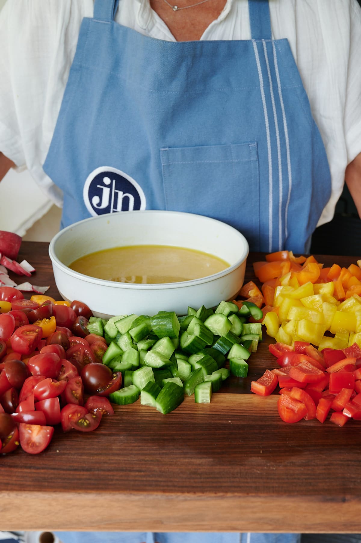 woman with blue apron holding a board with cut vegetables and wide bowl for salad dressing.