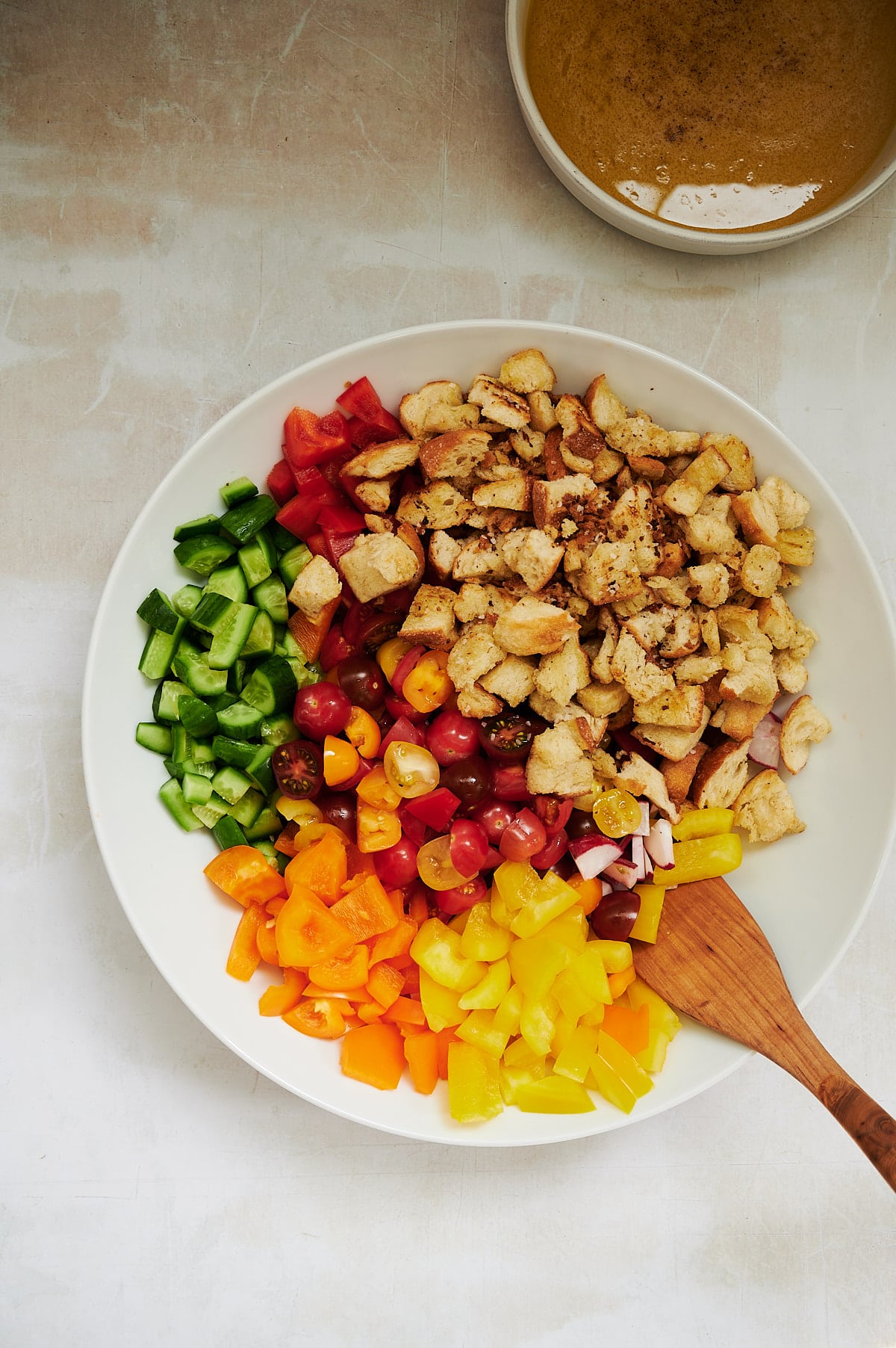 white bowl with cut up vegetables and cubed bread and salad dressing in a bowl.