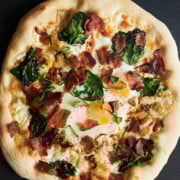 whole baked breakfast pizza with jammy eggs on a black pizza peel and small bowl of parmesan cheese.