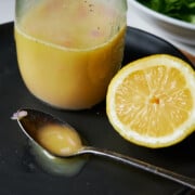 a lemon, a spoon with dressing and a clear bottle of lemon salad dressing sit on a black plate