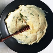 black bowl of mascarpone cheese dip topped with drizzle of honey, wooden honey dipper and sprig of thyme.