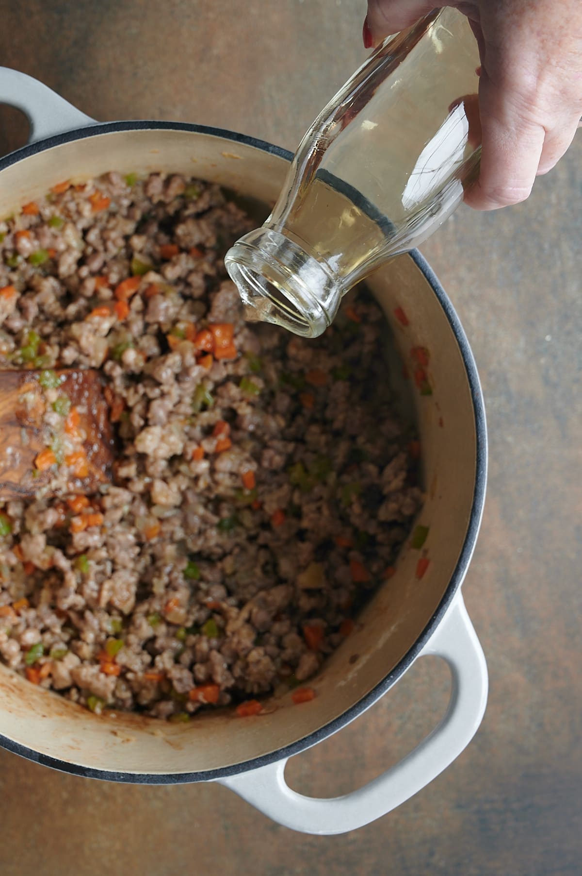 White wine being poured into browned ground meats in white Dutch oven.