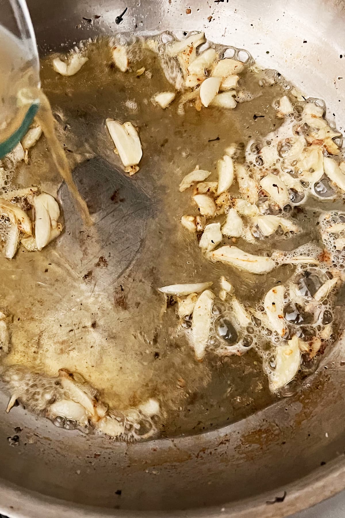 chicken broth being poured into skillet of oil and butter sauteing garlic
