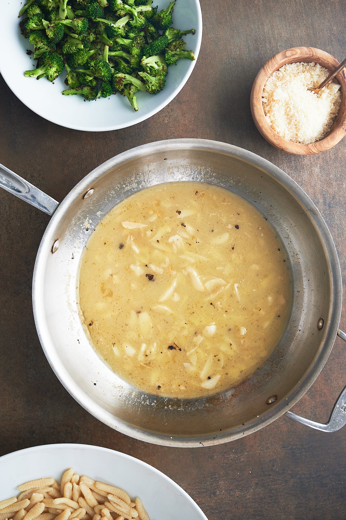 skillet of garlic in broth with bowls of broccoli, cavatelli and parmesan.