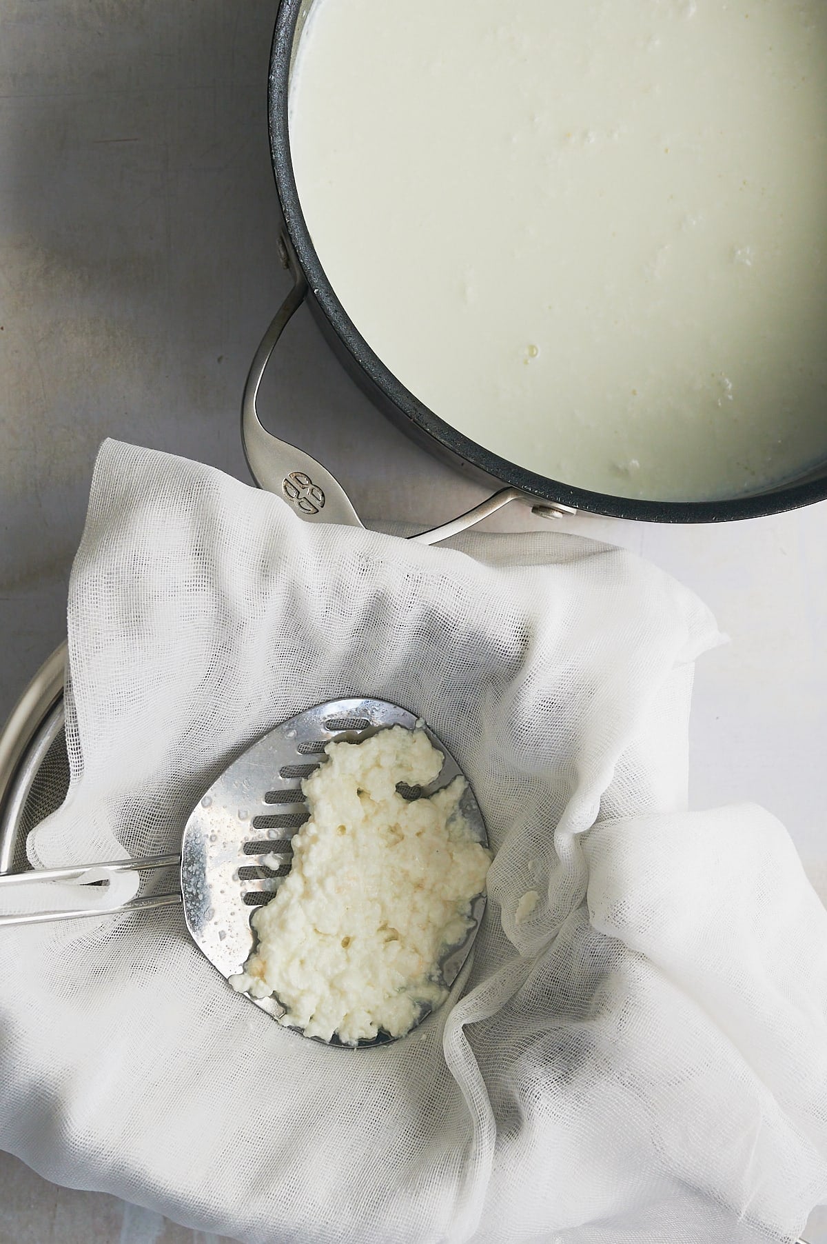 slotted spoon draining homemade ricotta cheese over cheesecloth with pot of cheese nearby.