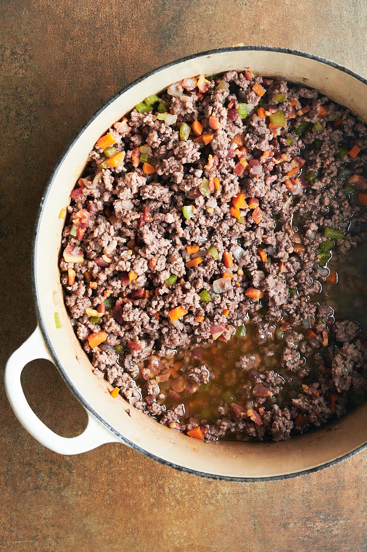 White pot of ground beef and pork with carrots, celery and onions with broth for bolognese sauce on a brown background.