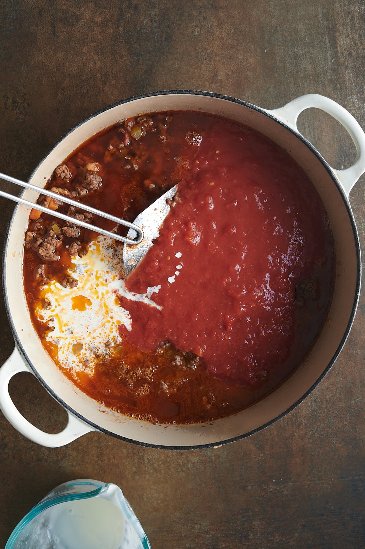 Cream added to bolognese cream sauce with tomatoes, ground meat and a silver strainer stirring all in a white dutch oven.
