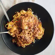 Black bowl of tagliatelle bolognese with cream being twirled with fork and a spoon on a white background with lace trimmed napkin and wooden bowl of parmesan.