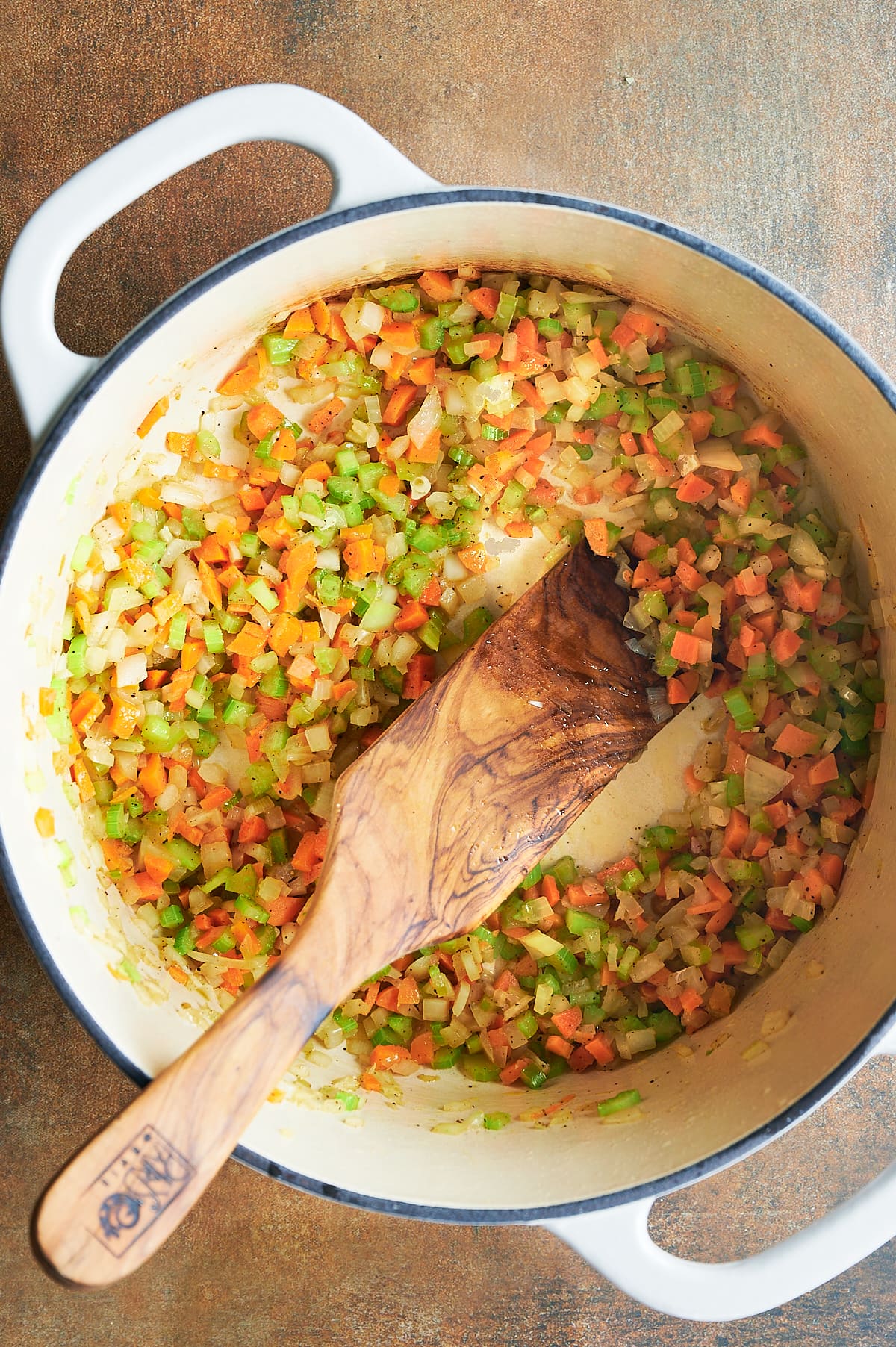 White pot of celery, onions and carrots being sauteed with a wooden paddle for bolognese sauce.