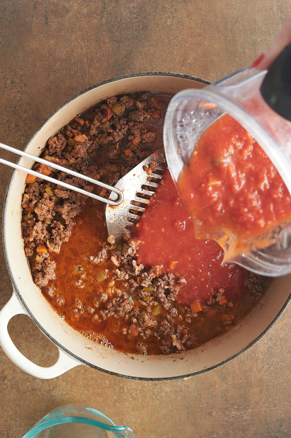 Tomatoes being poured from a blender into a white pot of ground beef and pork for Bolognese sauce with a silver strainer.