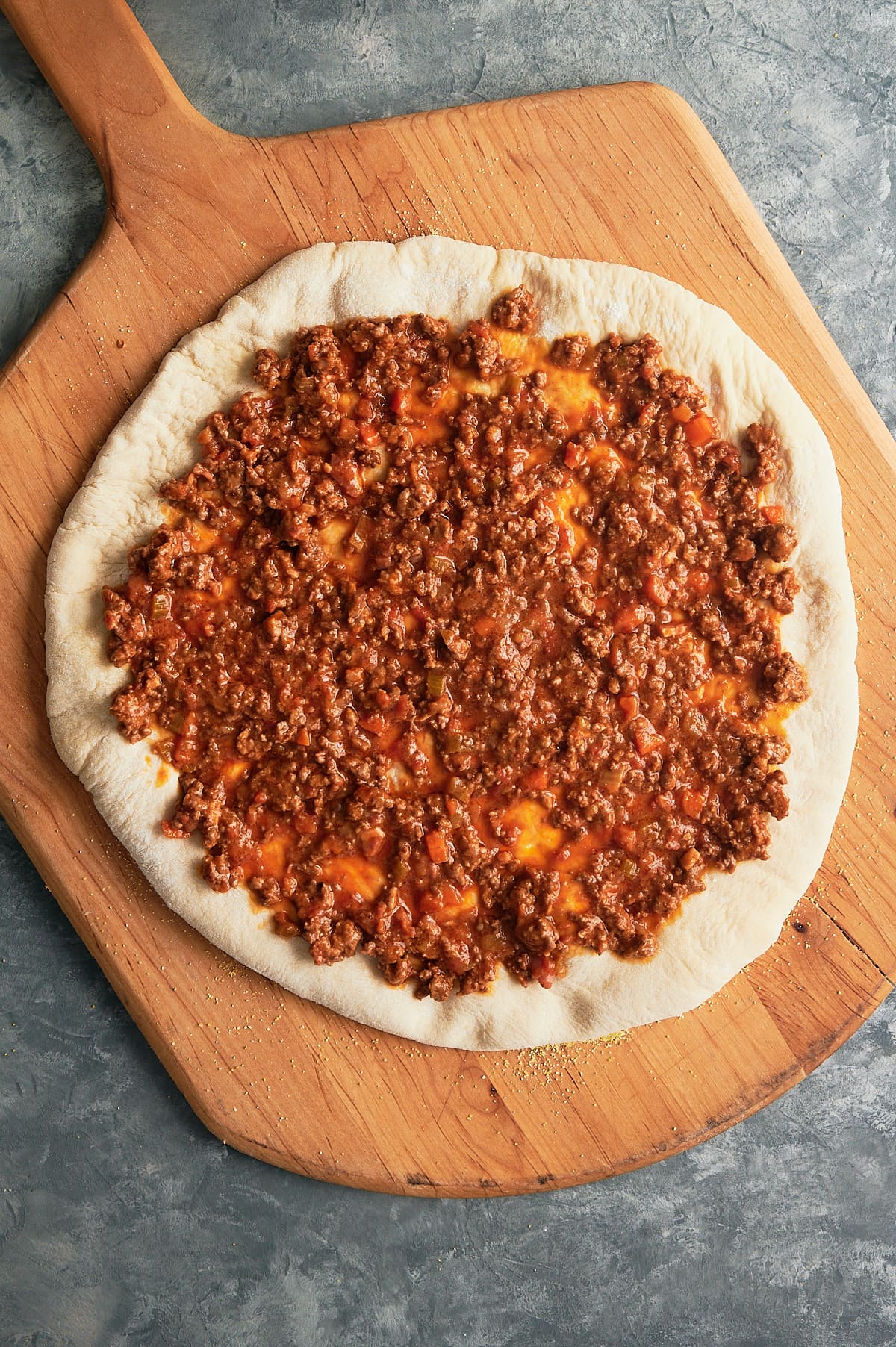 pizza crust topped with meat Bolognese sauce on wooden pizza peel.