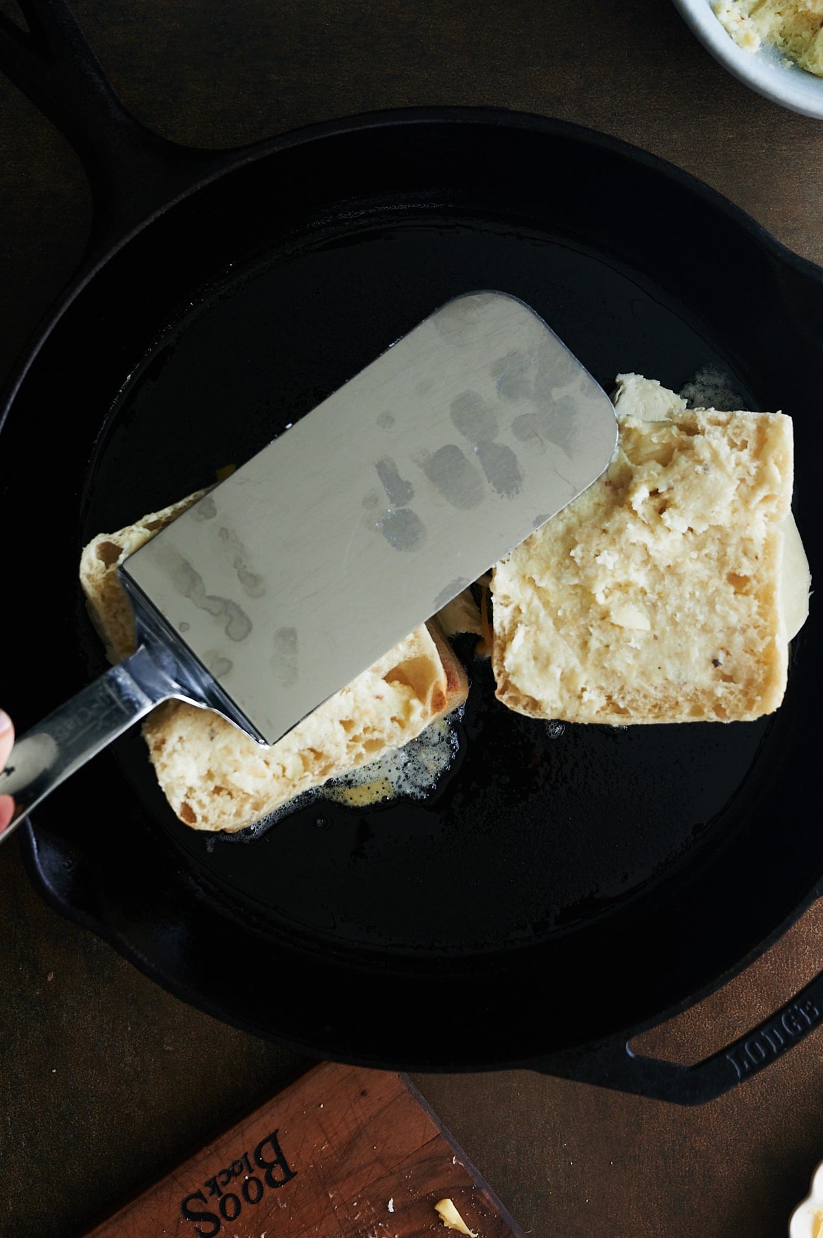 Spatula flattening ciabatta grilled cheese sandwiches in cast iron skillet.
