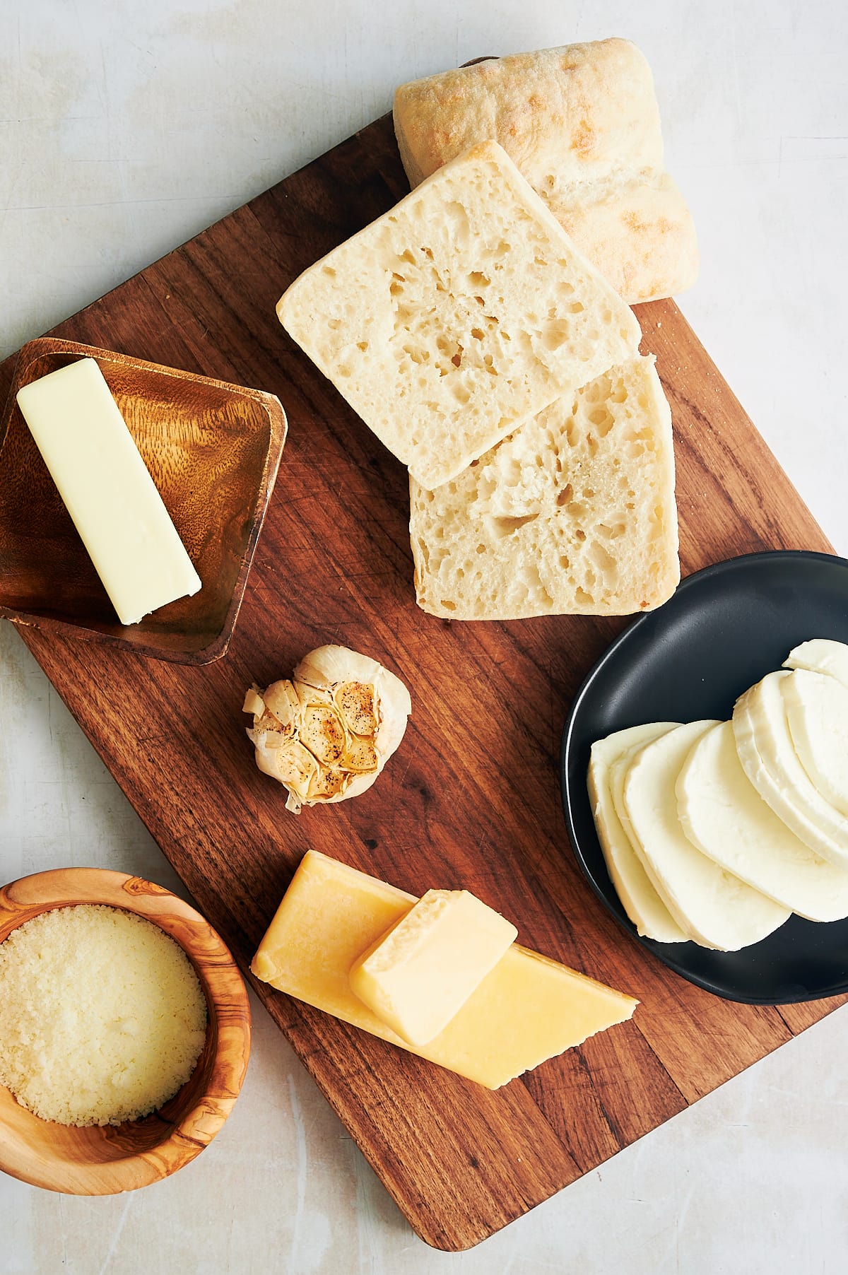 Wooden board with ciabatta rolls, garlic, fresh mozzarella and butter for grilled garlic cheese sandwiches.