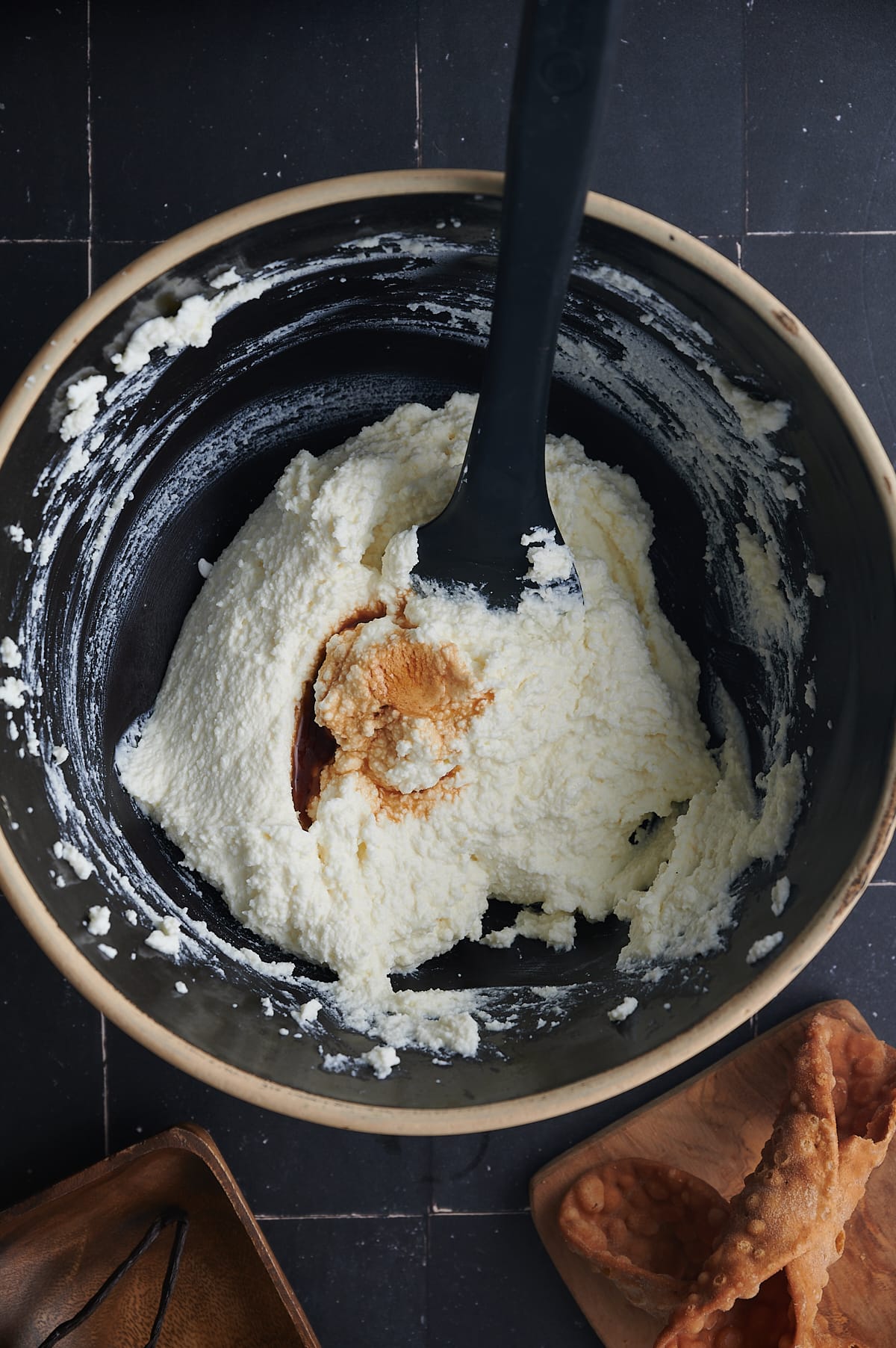 vanilla being mixed into cannoli filling with spatula in black bowl.