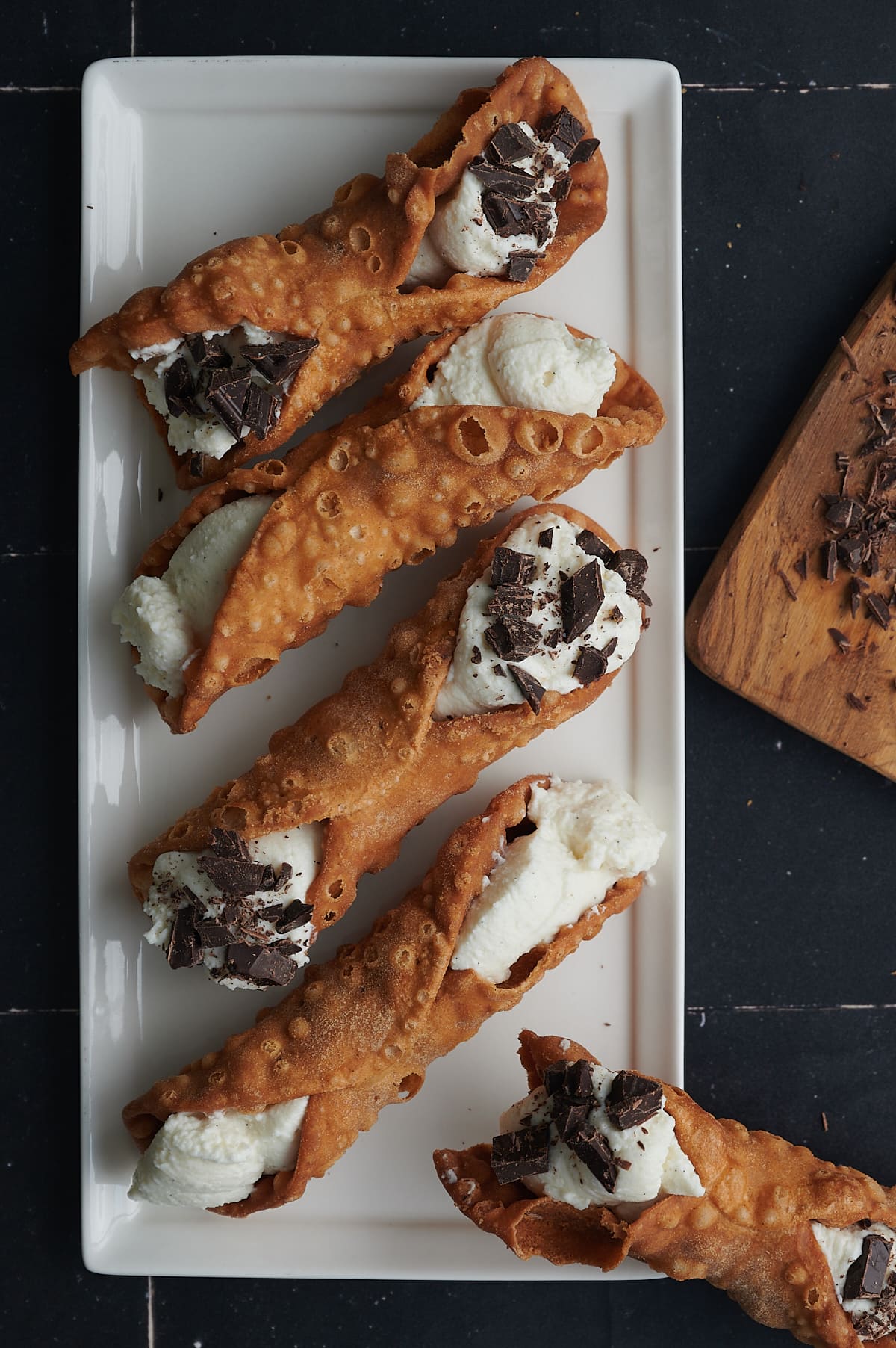 overhead of white tray of filled cannoli and cutting board of chopped chocolate.