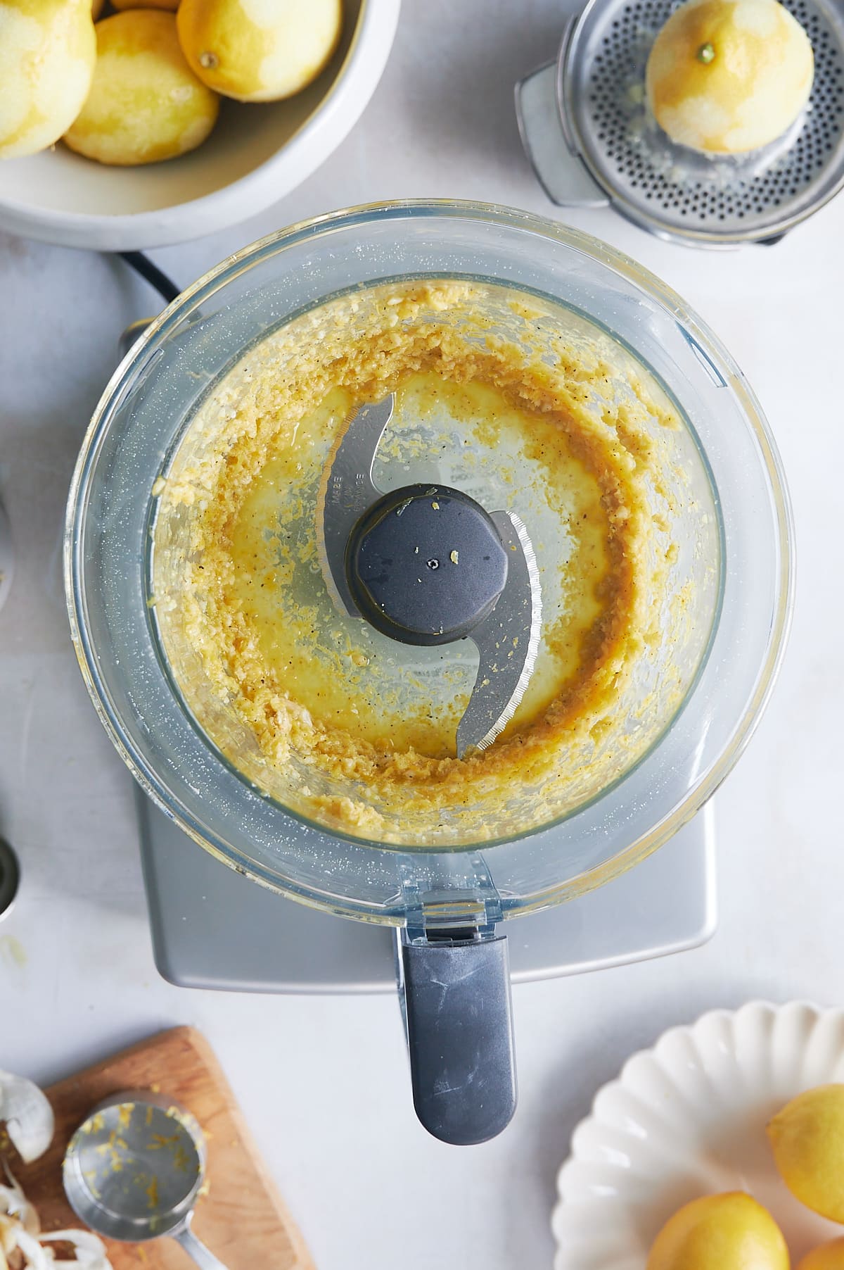 food processor with oil being added to lemon pesto with zested lemons nearby.