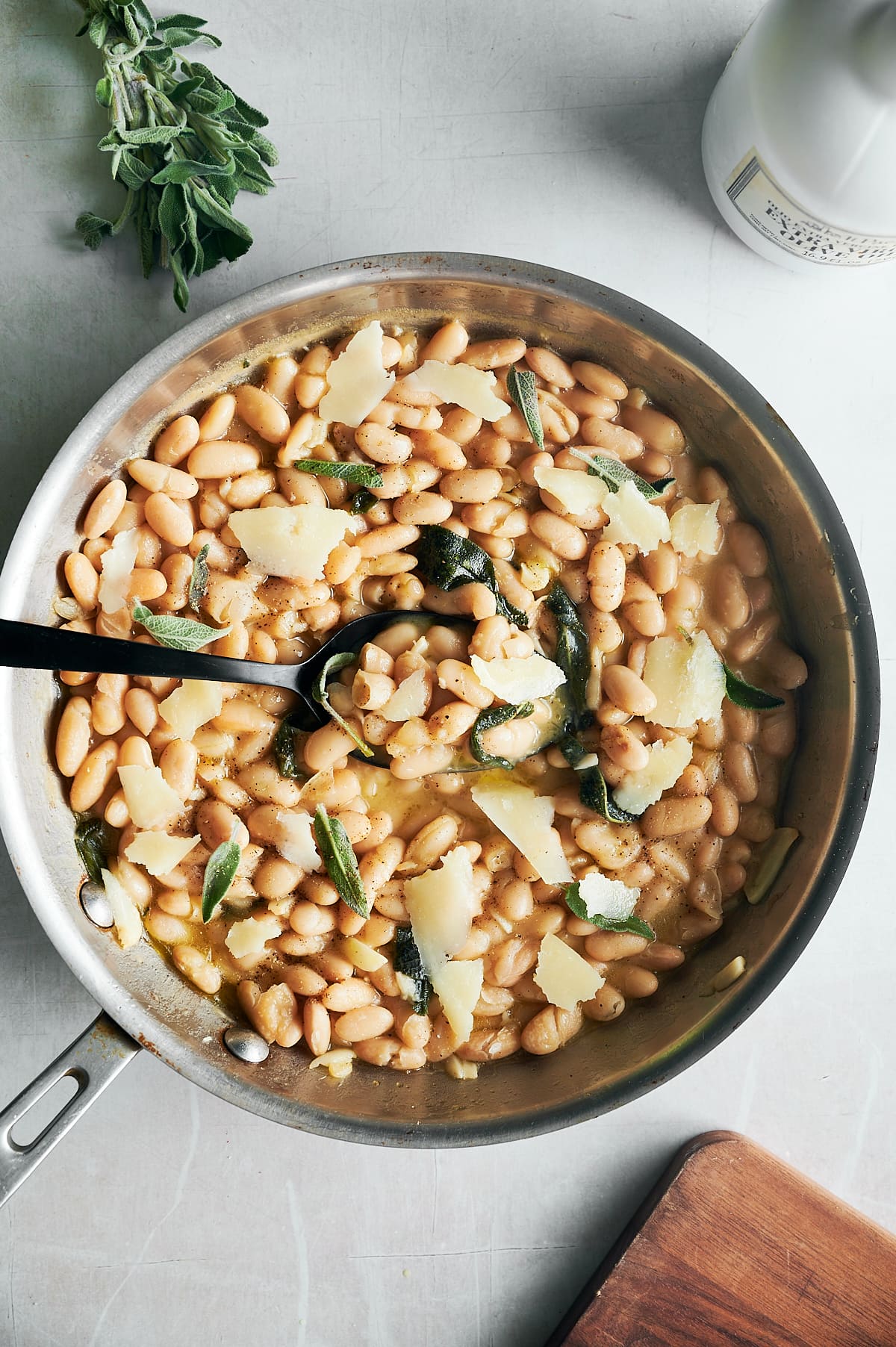skillet of a black spoon scooping up cannellini beans with sage and parmesan shards.