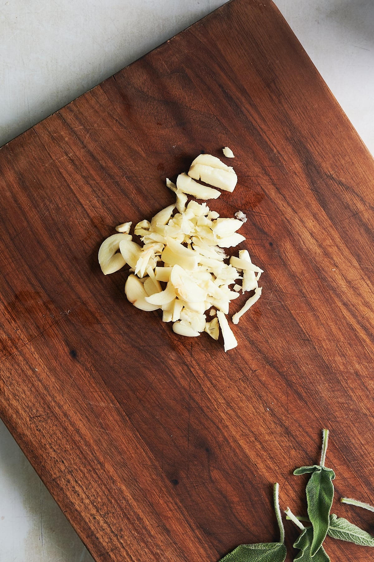 wooden cutting board of thinly sliced garlic with fresh sage leaves.