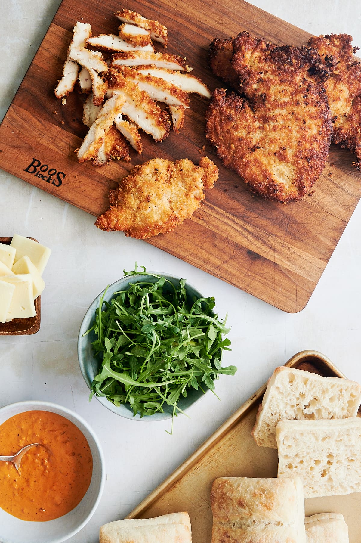 breaded chicken cutlets on a wooden board surrounded by bowls of arugula, vodka sauce and rolls for a chicken sandwich.
