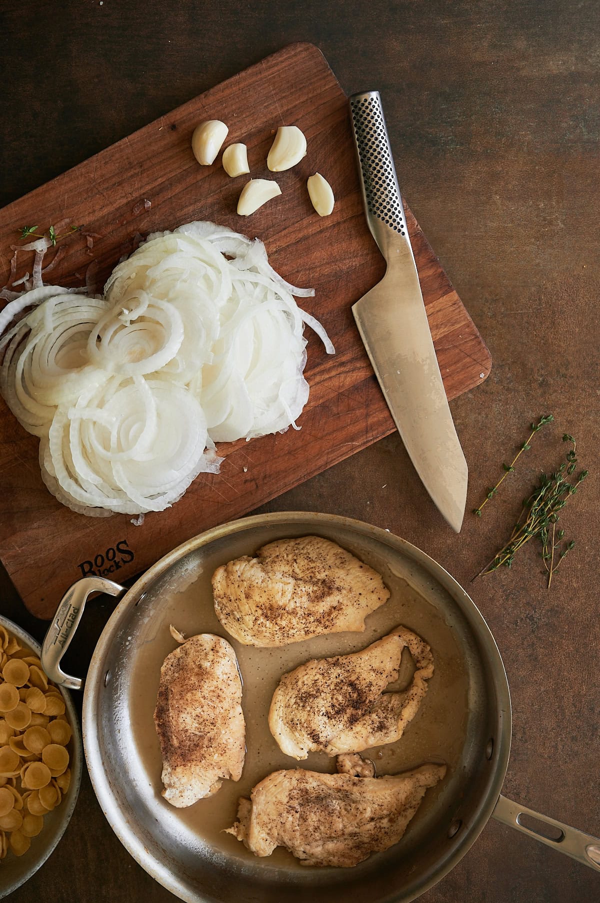 wooden board with knife and thinly sliced onions and pan of cooked chicken breasts.
