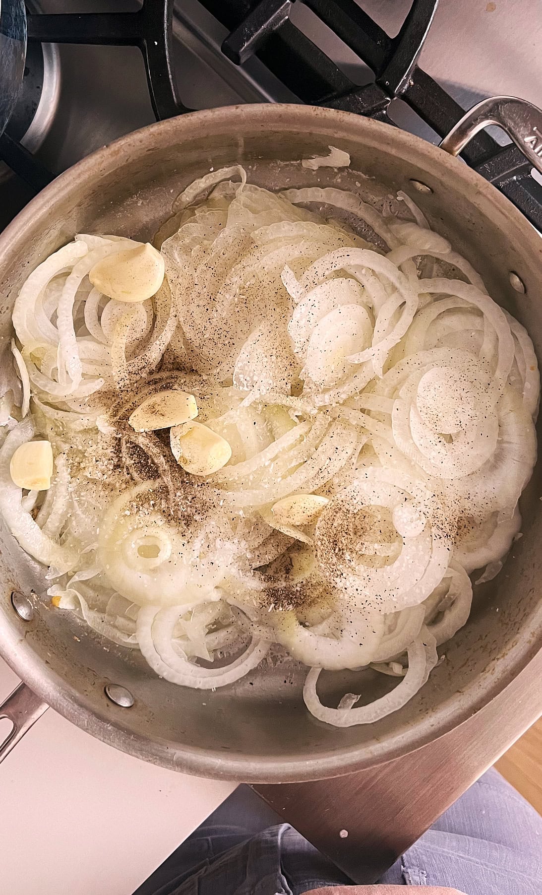 stainless pan of sliced onions with garlic cloves seasoned for caramelizing.