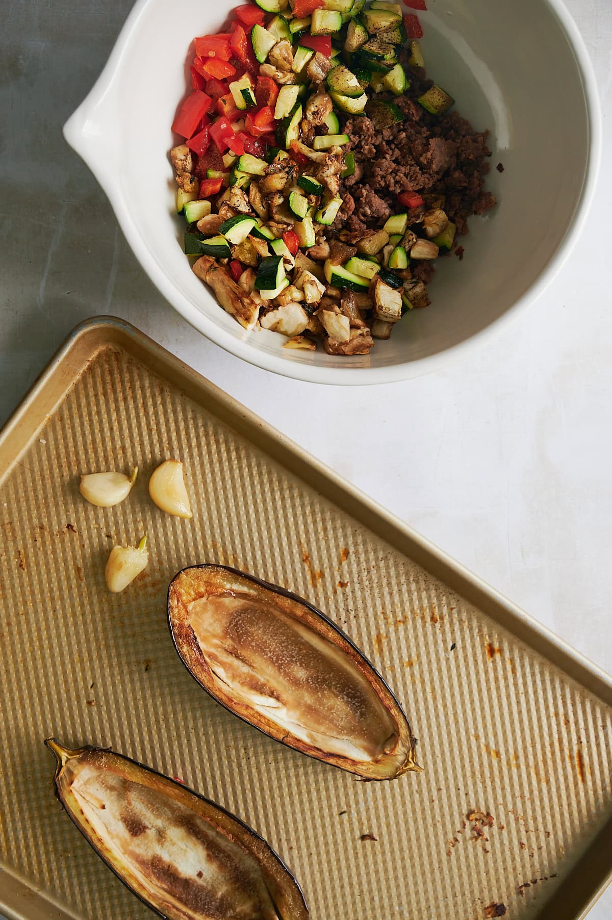 Roasted vegetables and cooked Italian sausage in white bowl near sheet pan of roasted eggplant halves and garlic cloves.