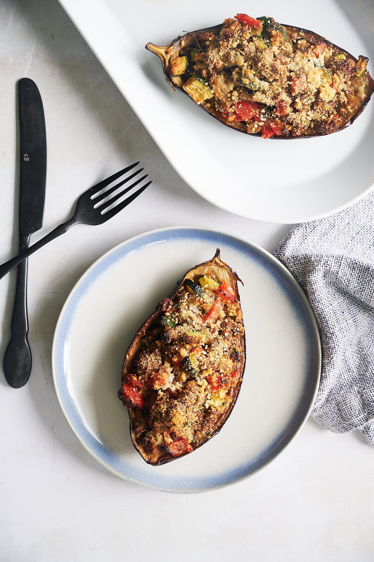 blue rimmed white plate with baked stuffed eggplant, black fork and knife and platter with other eggplant half.
