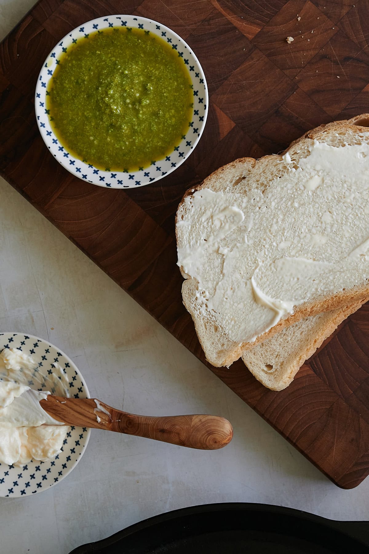 mayonnaise being spread onto slices of bread near a bowl of pesto sauce.