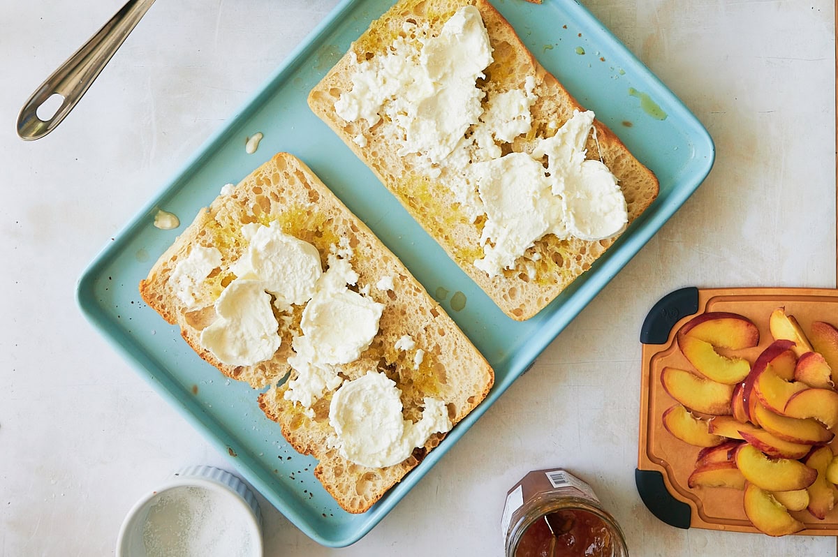 large toasted ciabatta bread spread with burrata on blue sheet pan with sliced peaches on board.