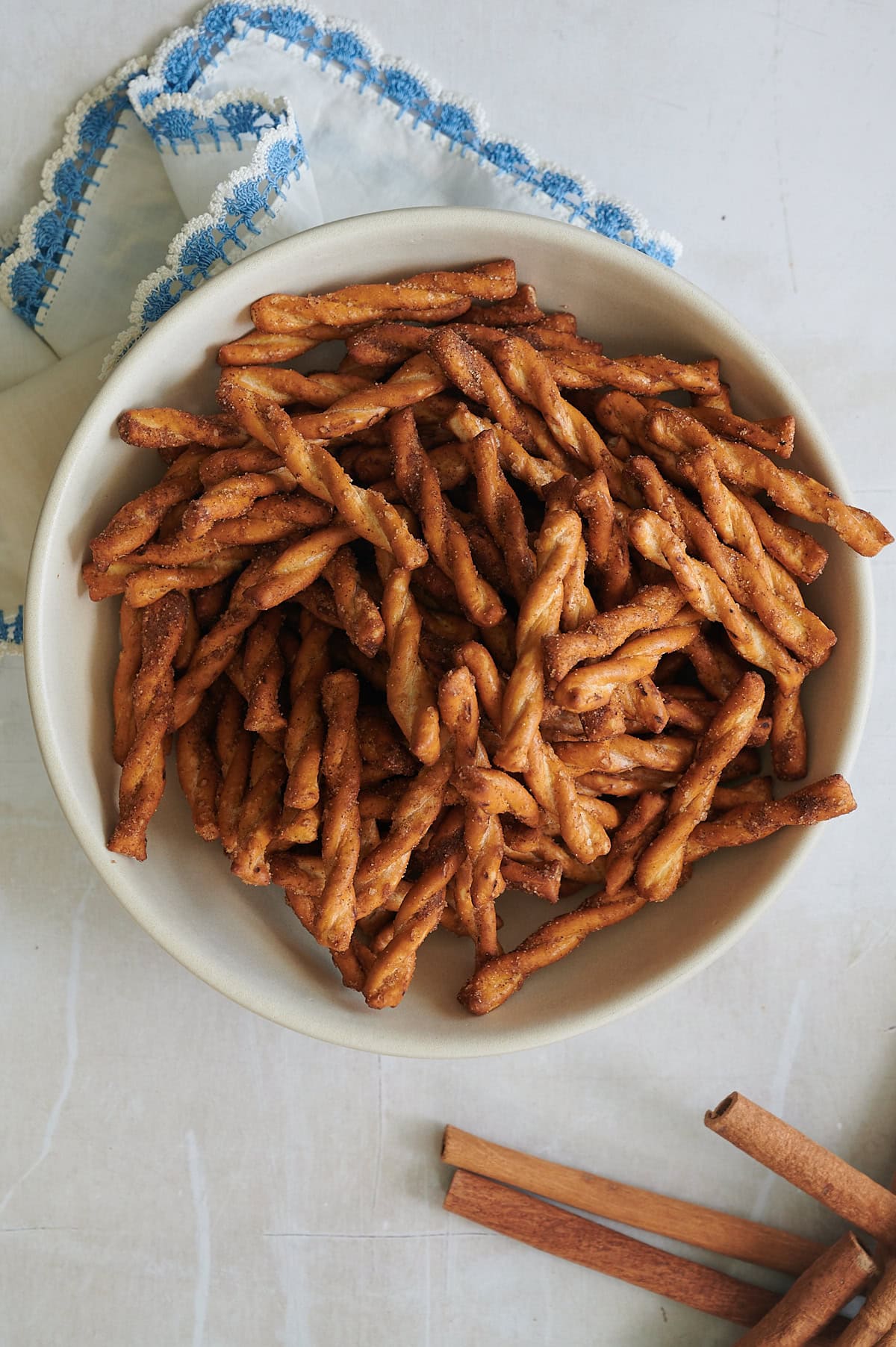 finished white bowl filled with cinnamon pretzels sitting on a white napkin with blue crocheted trim and cinnamon sticks nearby.